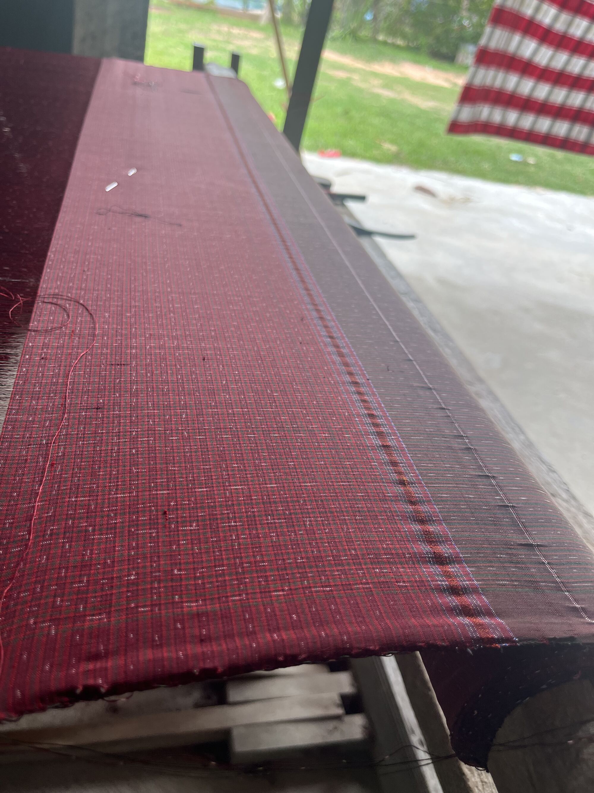 A length of deep red silk stretched on a wooden loom in the shade under the house, green yard beyond
